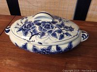 Side angled view of oval blue and white covered dish with floral Flow Blue pattern and dual side handles.