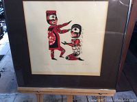 Framed silkscreen art showing two stylized Indigenous figures, one standing, one kneeling, with red, black, white colors, behind glass with matting and wood frame