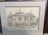 Framed art print of pencil sketch showing front and side view of a brick house with landscaping.