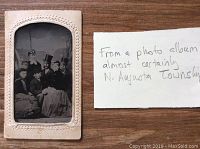 Tintype photo mounted on white card with embossed border, showing seven people in 1860s clothing.