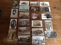 Overhead view of all 21 vintage postcards spread across a wooden table showing different black and white, sepia toned photographs including ships, people, houses, and street scenes.