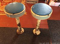 Full view of two antique cast enameled stools with blue cushioned seats and white bases with blue stripes, showing paint wear and rust.