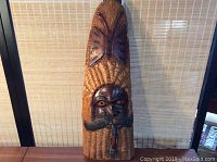 Full view of the carved wooden wall hanging standing upright on a flat surface against a woven backdrop.