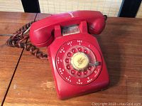 Front and top view showing the rotary dial, handset cradle, and coiled cord in red plastic with visible chip on cradle area.