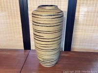 Front view of beige pottery vase with horizontal brown stripes on a wooden surface against a woven backdrop.