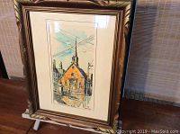 Full view of the framed watercolour artwork showing a Quebec church with a steeple, painted in ink and watercolour, placed on an easel.