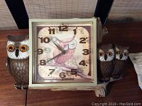 Vintage owl kitchen clock with three owls including a large owl on the left with bright orange eyes and two smaller owls on the right holding a square clock face in the center with a pink owl illustration and black numerals.