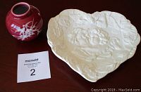 Photo showing small red ceramic vase and white heart-shaped ceramic plate with raised cupid relief, both on a wooden surface