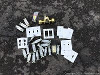 Photo showing an assortment of electrical outlets, switch plates, toggle switches in white, and a pair of brass door knobs laid out on pavement.