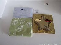 Green vinyl tablecloth in original packaging labeled 'Moda Essentials Leaf' shown with star-shaped gold decorative plate and textured square gold plate.