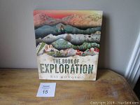 Front cover of 'The Book of Exploration' coffee table book by Ray Howgego on wooden surface with auction number card visible