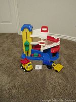 Fisher Price garage playset mostly blue, yellow, red, and white, standing on carpet with ramps and elevator visible, with two small toy trucks in front