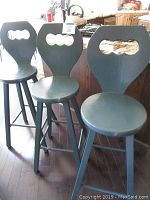 Three solid wood stools standing side by side on a dark wooden floor in front of a kitchen island. Each stool has a rounded seat, a wide back with a decorative cut-out, and four legs with horizontal supports. Painted in dark green.
