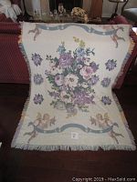 Full view of cream cotton woven blanket with floral bouquet and cherubs at corners, displayed over sofa back.