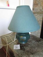Photo of ceramic lamp with green fabric shade sitting on stone surface with power cord visible, showing small chips on ceramic base and stain on shade.