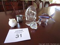 Overview of the six items including ceramic pig, metal shot glass, glass sheep block, blue glass fish, glass beaver, and glass elephant sculpture on wooden table.