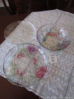 Three glass serving pieces on lace tablecloth, including two platters and one bowl with painted floral designs.