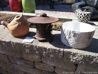 Photo showing three outdoor decorative items placed on a stone ledge in sunlight: a terracotta clay bird, a cast iron candle holder with a lid, and a white textured ceramic planter.