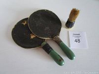 Two vintage ping pong paddles with visible wear on rubber surfaces and black tape on handles alongside a small Bakelite shaving brush with a dark handle and natural bristles