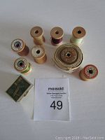Top view of lot including seven wooden thread spools with varying thread colors, a coiled tape measure in a round vintage fabric trinket box, and a small green box of sewing needles.