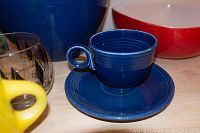 Photo showing close up of blue Fiesta cup on matching saucer next to glassware and partial view of ceramic bowls.