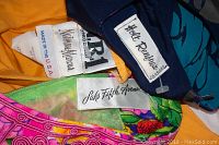 Close-up photo showing a cluster of labels from various dresses including 'Saks Fifth Avenue', 'Hot Reflections', 'Ann Gordon by Housecoats Inc.' and others.