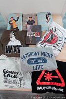 Full view of the comedy-themed shirts, Louis CK DVD and comedy books on a light wood floor.