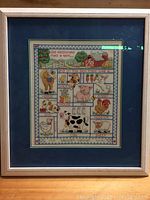 Full view of the framed needlepoint artwork showing entire piece with white frame and blue mat, featuring farm animals and barn scene.