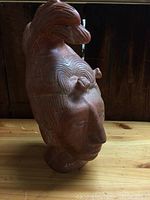 Side view of the reddish-brown clay bust showing detailed swirling carved patterns on the head and face profile.