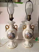 Full view of the pair of porcelain table lamps showing overall shape, gold accents, painted scenes.