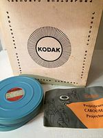 Beige Kodak box with black Kodak logo and text 'Projecteur Carousel' alongside one blue film reel with a red label and the projector manual.