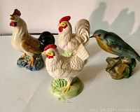 Photo of four ceramic bird figurines including three roosters and one blue bird on a white surface with shadows visible behind them.