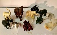 Twelve elephant figurines arranged on a flat surface with varied colors and materials visible, including dark wood, black resin, cream and white stone.