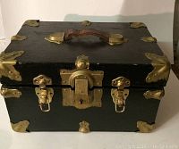 Front view of the closed miniature trunk showing black exterior, brass hardware, locks, and leather handle.
