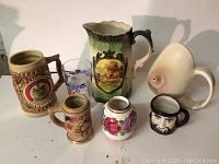 Seven-piece dish lot including two pitchers, two beer steins, a decorative glass, a small painted vase, and a face mug photographed on white surface.