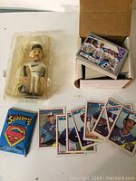Wide view of the bobblehead packaged on the left and a box of assorted sports cards on the right including Blue Jays baseball cards and a Superman II card pack.