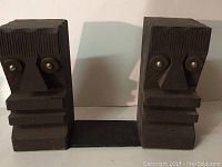 Front view of both wooden bookends featuring carved abstract faces and metal accents for eyes, connected by a black base.