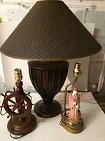 Full shot of the lamp with urn-shaped base and large brown textured shade, next to a wooden ship wheel lamp base and the fishing man sculptural lamp.