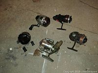 Photo of five assorted fishing reels placed on the ground with visible wear and dirt.