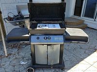 Full view of Broil Mate barbecue grill with lid open showing black and stainless steel construction, fold-down side shelves, front doors, and manual on grill grate.