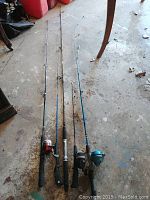 Photo showing four fishing rods laid side by side with reels attached, captured from above at an angle on a concrete floor.