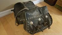 Side view of vintage black leather motorcycle saddle bags showing studded detail and decorative conchos with tassels on front flap