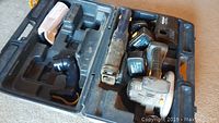 View of tools and accessories placed inside the open hard plastic carrying case. Includes two cordless saws (reciprocating and circular), charger, and batteries arranged in compartments.