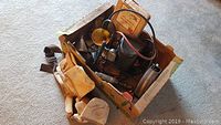 Box containing multiple tools including a vintage work light, gauges, hoses, and other tools in a cardboard box.