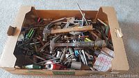 Photo showing a large cardboard box filled with assorted hand tools including hammers, screwdrivers, wrenches and other items scattered inside.