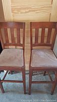 Two wooden bar stools with vertical slatted backs and cushioned beige seats placed side by side against a wooden wall and carpeted floor.