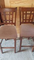 Photo showing full front view of two wooden bar stools with lattice backrests and brown fabric cushioned seats on carpeted floor.