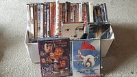 Box of VHS tapes and DVDs showing titles such as The Lord of the Rings and Airplane in front.
