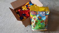 Photo of an open cardboard box containing assorted loose Mega Bloks blocks and a sealed yellow bag of Mega Bloks 160-piece set placed in front