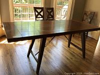 Photo showing the table in front of large windows with three visible chairs (not included). Shows wood grain detail and sturdy angled legs.